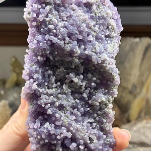 May include: A close-up of a Grape Agate specimen. The surface is covered in small, round, lavender-colored formations, with hints of green. The overall texture is bumpy and clustered, resembling a bunch of grapes. The stone is held in a hand.