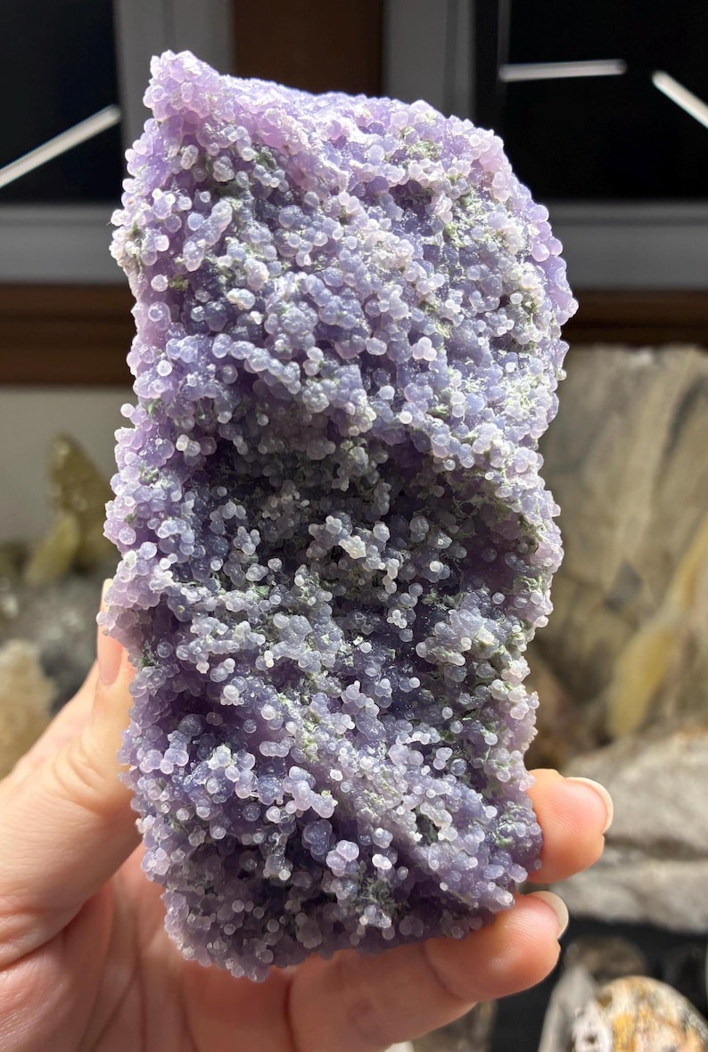 May include: A close-up of a Grape Agate specimen. The surface is covered in small, round, lavender-colored formations, with hints of green. The overall texture is bumpy and clustered, resembling a bunch of grapes. The stone is held in a hand.