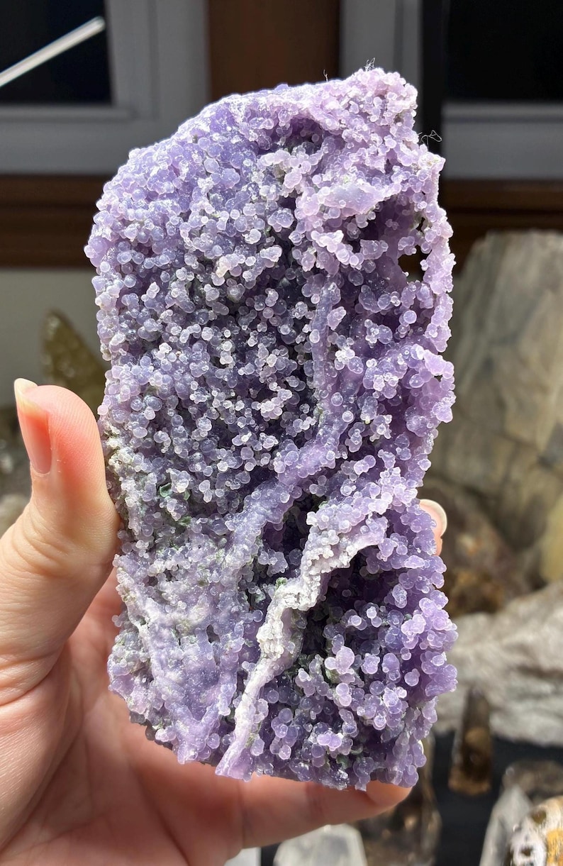 May include: A close-up of a grape agate specimen. The stone features a bubbly, botryoidal texture in shades of lavender and purple. The surface has a crystalline appearance, with a few areas of white and green.