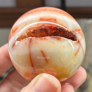 May include: A polished, spherical gemstone with a mix of orange, white, and brown hues. The stone has a crack running through it, revealing a textured interior. The overall appearance is smooth and glossy, with visible color variations.