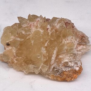 May include: A cluster of light amber-colored crystals with a rough, textured surface. The crystals have a layered appearance with hints of white and reddish-brown hues. The overall shape is irregular and natural, suggesting a mineral specimen.