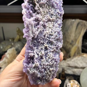 May include: A close-up of a grape agate specimen. The crystal features a clustered, rounded formation with a light purple hue and areas of gray and white. The surface has a bumpy texture.