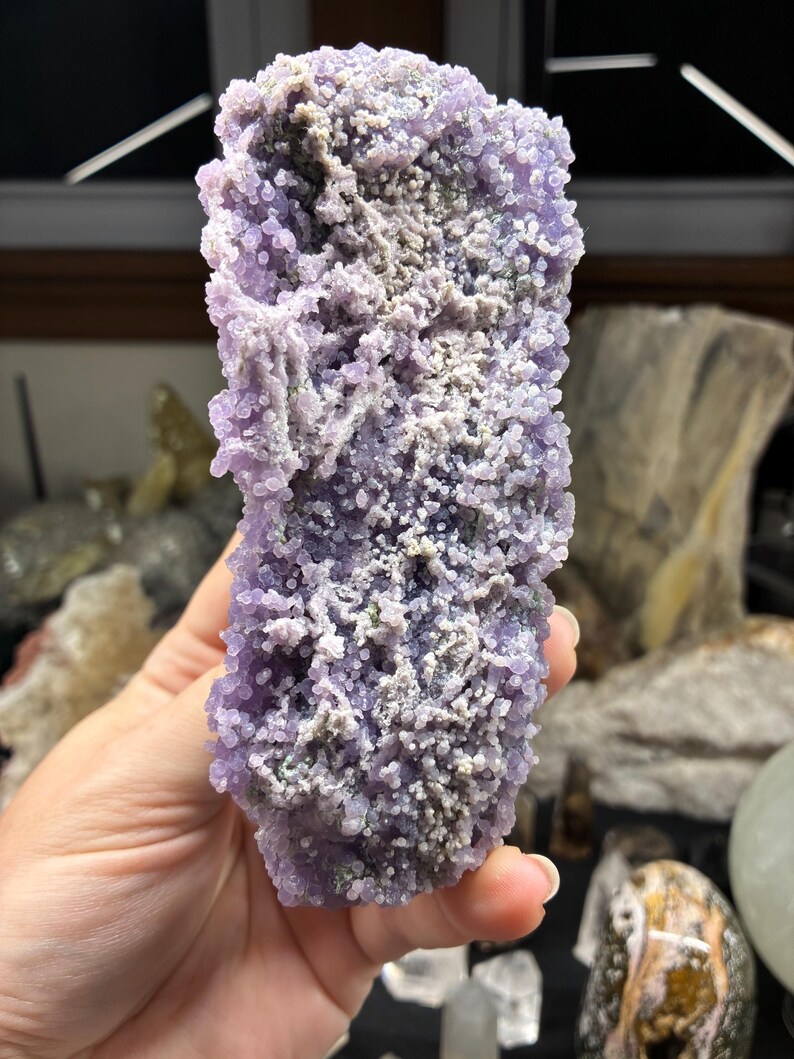 May include: A close-up of a grape agate specimen. The crystal features a clustered, rounded formation with a light purple hue and areas of gray and white. The surface has a bumpy texture.