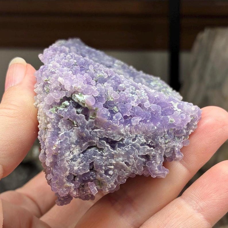May include: A close-up of a piece of grape agate, a unique mineral formation. The surface is covered in small, spherical, lavender-colored nodules, with hints of green. The overall texture is bumpy and irregular, showcasing the natural beauty of the stone.