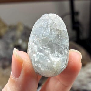 May include: A pale blue, oval-shaped crystal held between fingers. The crystal has a rough, fractured surface, catching the light and revealing internal structures. The overall appearance is translucent, with subtle variations in color and texture.