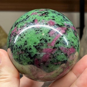 May include: A polished sphere of zoisite with ruby inclusions. The gemstone has a vibrant green base colour with dark black speckles and streaks of deep pink ruby. The sphere is held in a hand, showcasing its smooth, reflective surface.