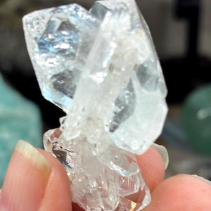 May include: A cluster of clear, faceted quartz crystals. The crystals have a translucent, icy appearance with visible internal structures. The quartz is held in a hand, showcasing its natural, geometric formations. The overall effect is one of natural beauty and geological interest.