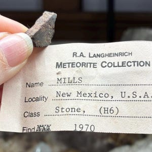 May include: A small, dark-colored meteorite fragment held near a label from the R.A. Langheinrich Meteorite Collection. The label indicates the meteorite's name is MILLS, from New Mexico, USA, and was found in 1970.
