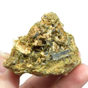 May include: A rough, natural mineral specimen featuring a mix of green, brown, and beige hues. The stone has a textured surface with visible crystalline structures and is held in a hand.