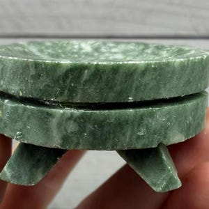 May include: A small, green stone sculpture resembling a miniature table. The piece is crafted from a mottled green stone, featuring a circular top and base, connected by a central section. Three small legs support the structure.