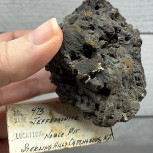 May include: A dark, porous, and textured mineral specimen identified as Jeffersonite. Attached is a rectangular paper label with handwritten text: No. 473, Name Jeffersonite, Location Noble Pit, Sterling Hill, Ogdensburg, N.J.
