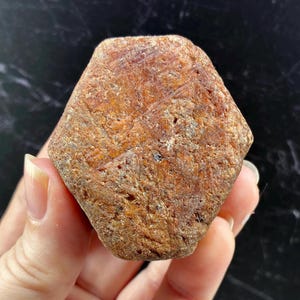 May include: A raw, hexagonal-shaped mineral specimen with a rough, textured surface. The stone displays earthy tones of reddish-brown, rust, and ochre, featuring natural striations and small dark inclusions across its uneven, weathered exterior.
