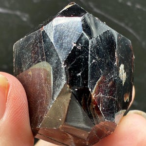 May include: A faceted, dark reddish-brown garnet crystal with a polished, geometric surface. The stone features multiple sharp, angular planes that reflect light, revealing deep, translucent crimson tones within its dense, dark structure.