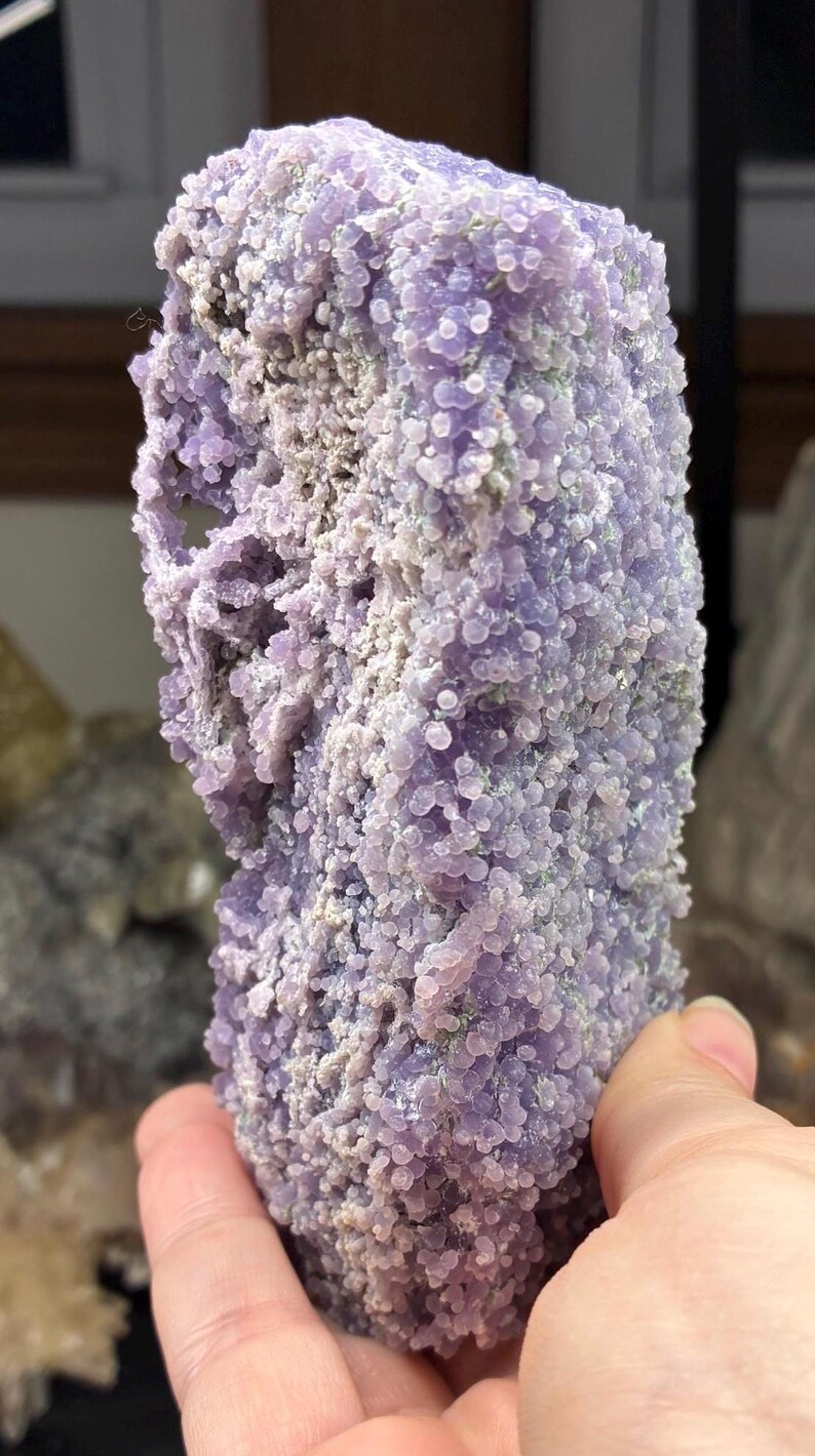 May include: A large, raw, grape agate specimen. The surface is covered in small, round, lavender-colored botryoidal formations. The stone has a rough, textured appearance with areas of gray and white.