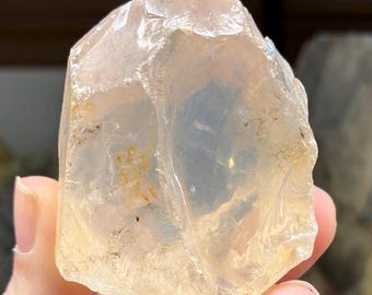May include: A translucent, rough-cut gemstone with a pale yellow and clear color. The stone has a crystalline structure and is held in a hand. The gemstone is likely a raw mineral specimen.