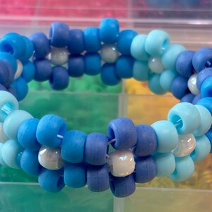 Puede incluir: Un brazalete de cuentas multicolores con cuentas azules, azul claro y blancas. El brazalete está hecho de cuentas pequeñas y redondas que están enhebradas en una cuerda.