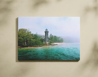 May include: A canvas print featuring a coastal scene with a lighthouse. The lighthouse is a weathered gray structure with a tall tower, set against a backdrop of lush green trees and a misty sky. The water is a vibrant turquoise.
