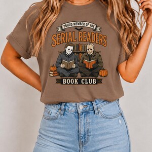May include: A brown short-sleeve t-shirt featuring a graphic print of two masked figures sitting on the floor reading books in front of a bookshelf. The text reads PROUD MEMBER OF THE SERIAL READERS BOOK CLUB with orange and black accents.
