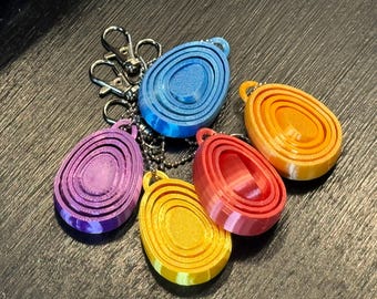 Könnte beinhalten: Eine Sammlung farbenfroher, 3D-gedruckter Schlüsselanhänger in Blau, Lila, Rot, Gelb und Orange. Jeder Schlüsselanhänger hat eine ovale Form mit konzentrischen Ringen und einen Metallverschluss. Die Schlüsselanhänger sind auf einer dunklen Oberfläche angeordnet.
