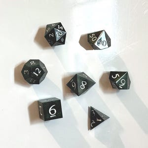 May include: Seven black polyhedral dice with white numbering are arranged on a white surface. The dice include a D20, D100, D12, D8, D6, D4, and a D10. The dice are for tabletop role-playing games.
