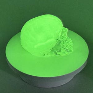 May include: A luminous green skull rests on a circular green base. The skull's features, including eye sockets and teeth, are clearly defined. The base is supported by a grey, cylindrical pedestal, creating a contrast against the dark green backdrop.