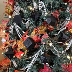 以下が含まれることがあります： さまざまな黒い多面体サイコロのオーナメントで飾られたクリスマスツリー。ツリーは白いビーズガーランド、秋の葉、小さな白いライトで飾られています。サイコロには番号が付けられており、さまざまな幾何学的形状をしています。