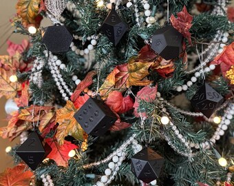 Puede incluir: Un árbol de Navidad decorado con varios adornos de dados poliédricos negros. El árbol está adornado con guirnaldas de cuentas blancas, hojas de otoño y pequeñas luces blancas. Los dados están numerados y tienen diferentes formas geométricas.