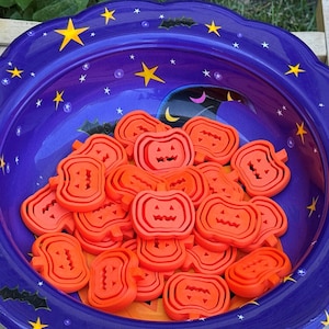 May include: A purple Halloween candy bowl with a scalloped edge, decorated with yellow stars and bats. The bowl is filled with orange pumpkin-shaped candies, perfect for trick-or-treaters.