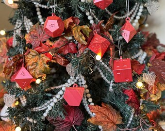 Puede incluir: Un árbol de Navidad decorado con adornos rojos en forma de dados. Los adornos cuelgan del árbol con una cuerda blanca. El árbol está decorado con hojas de otoño, cuentas blancas y pequeñas luces. Los dados tienen números.