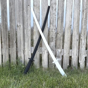 May include: Two katana swords, one black and one white, lean against a weathered wooden fence. The swords are crossed, with their handles and blades visible. The scene is set outdoors with green grass. The swords are approximately 100 cm in length.