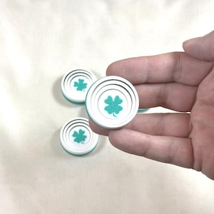 May include: White and teal round game pieces with a four-leaf clover design. The pieces have a white center with a teal clover and concentric circles, and a teal edge. Several pieces are visible, with one held in a hand.