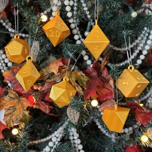 May include: A collection of gold-colored polyhedral dice ornaments hanging on a Christmas tree. The ornaments are various shapes, including d4, d6, d8, d10, d12, and d20 dice. The tree is decorated with white beads and autumn leaves.