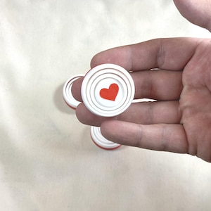 May include: White and red game pieces with a concentric circle design and a red heart in the center. The game pieces are round and appear to be made of plastic. Several pieces are visible.