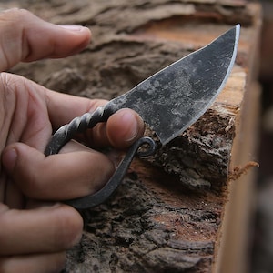 May include: A small, hand-forged knife with a curved blade and a twisted handle. The blade is dark grey, and the handle is a matching dark metal. The knife is held against a rough, brown wooden surface.
