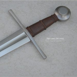 Peut inclure: Une épée médiévale avec une lame en acier poli et une poignée recouverte de cuir marron. La garde comporte une garde transversale et un pommeau arrondi. L'épée est présentée sur un fond neutre.