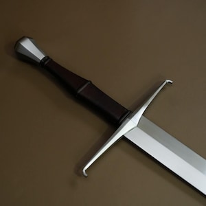Peut inclure: Une épée de style médiéval avec une lame en acier poli et un manche gainé de cuir marron foncé. La garde et le pommeau sont argentés. L'épée est présentée sur un fond marron.