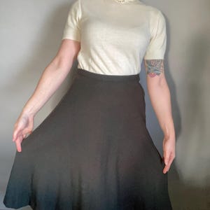 May include: A vintage outfit featuring a cream-colored short-sleeve sweater with a decorative collar and a dark charcoal-colored flared skirt. The skirt has a flowing silhouette. The outfit is suitable for a retro or classic style.