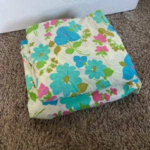 May include: Folded white bed sheet set with a floral pattern. The design features large turquoise and blue flowers with pink accents and green leaves. The sheets are folded on a brown carpet.
