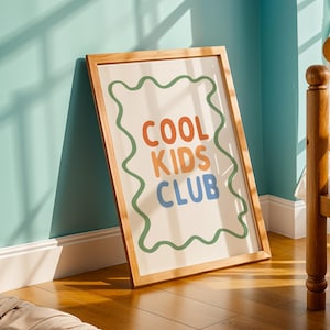 May include: A framed print with the words "COOL KIDS CLUB" in orange, light orange, and blue, outlined by a green wavy border. The print is in a light wood frame and leans against a teal wall.