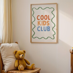 May include: A framed poster with the words "COOL KIDS CLUB" in orange, blue, and orange, with a green wavy border. A brown teddy bear sits on a wooden chair with a cream-colored pillow. A small wooden shelf holds two more teddy bears.