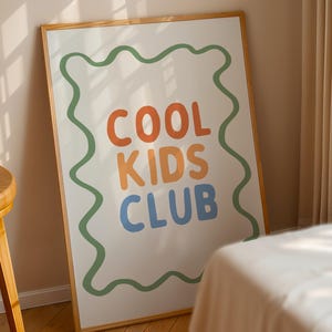 May include: A framed poster with the words "COOL KIDS CLUB" in orange, yellow, and blue, outlined by a green wavy border. The poster is displayed against a beige wall and a wooden frame.