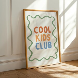 May include: A framed art print with the words "COOL KIDS CLUB" in orange, blue, and orange letters. The text is inside a green wavy border on a white background. The frame is a light brown color, and the print is leaning against a white wall.