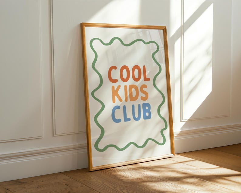 May include: A framed art print with the words "COOL KIDS CLUB" in orange, blue, and orange letters. The text is inside a green wavy border on a white background. The frame is a light brown color, and the print is leaning against a white wall.