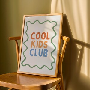 May include: A framed print with the words "COOL KIDS CLUB" in orange, blue, and green. The text is centered on a white background, framed by a wavy green border. The print is displayed on a wooden chair, with sunlight casting shadows.