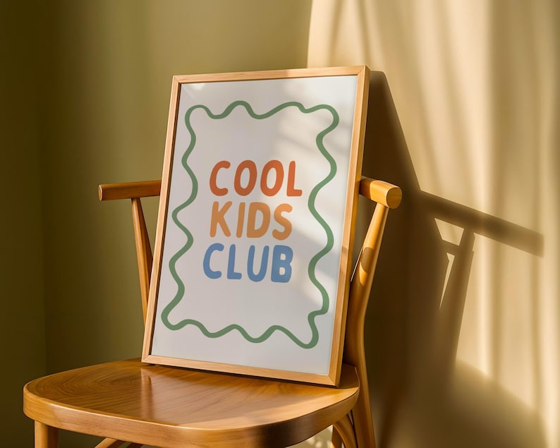 May include: A framed print with the words "COOL KIDS CLUB" in orange, blue, and green. The text is centered on a white background, framed by a wavy green border. The print is displayed on a wooden chair, with sunlight casting shadows.