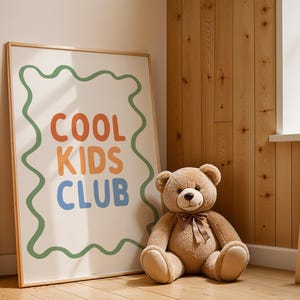 May include: A framed poster with the words "COOL KIDS CLUB" in orange, blue, and green, with a green wavy border. A large brown teddy bear with a brown ribbon sits next to the poster. The background is a wooden wall.