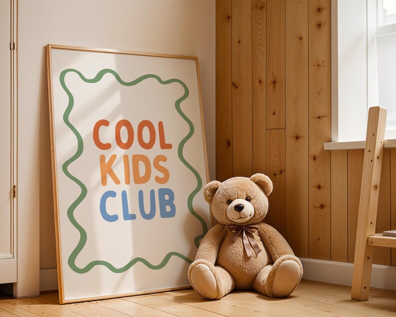 May include: A framed poster with the words "COOL KIDS CLUB" in orange, blue, and green, with a green wavy border. A large brown teddy bear with a brown ribbon sits next to the poster. The background is a wooden wall.