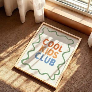 May include: A framed art print with the words "COOL KIDS CLUB" in orange, blue, and green. The text is on a white background with a green wavy border, set in a light wood frame. The artwork is displayed on a brown carpet.
