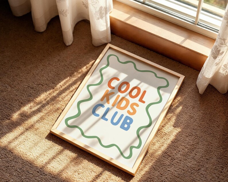 May include: A framed art print with the words "COOL KIDS CLUB" in orange, blue, and green. The text is on a white background with a green wavy border, set in a light wood frame. The artwork is displayed on a brown carpet.