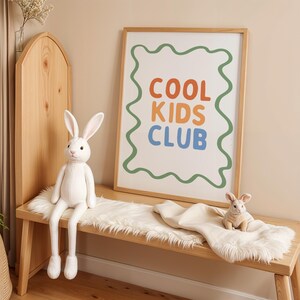 May include: A wooden bench with a white faux fur throw and two stuffed bunny rabbits. A framed print with the words "COOL KIDS CLUB" in orange, blue, and green is on the wall. The room has a neutral color palette.